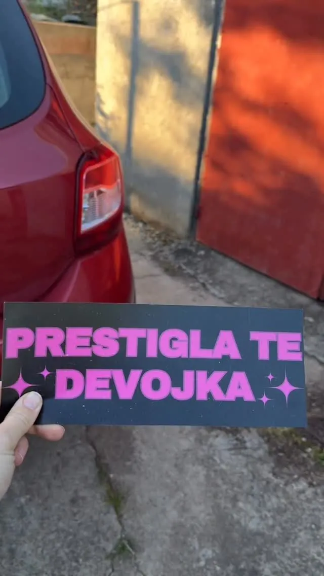 Ruka drži tablu „Prestigla te devojka” pored crvenog auta